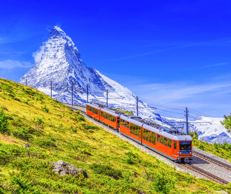 An orange train travels along a mountainside track with a snow-covered peak in the background under a clear blue sky.