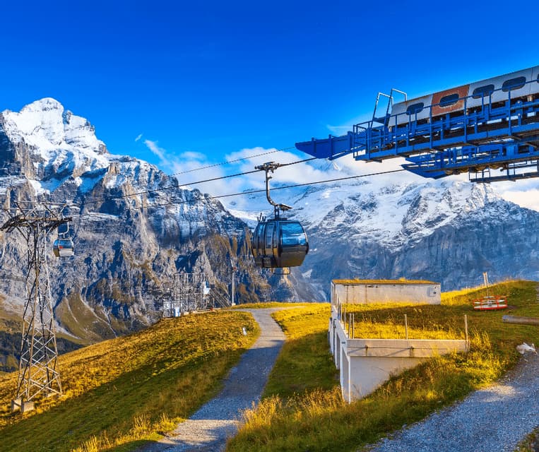 A gondola travels on a cable over a grassy mountainside, with snow-covered peaks visible in the background under a blue sky.