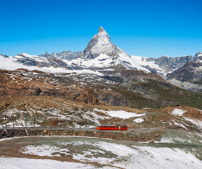 A red train travels on tracks along a snowy mountainside, with a large, pointed snow-capped peak against a clear blue sky.