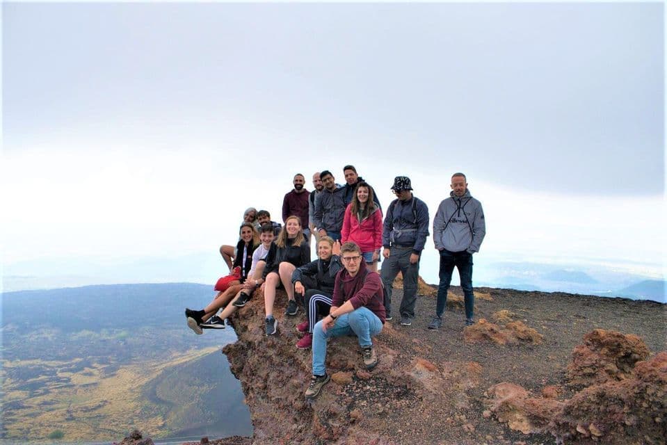 A WeRoad group trip posing together on a rocky, volcanic outcrop overlooking a vast landscape under a cloudy sky.