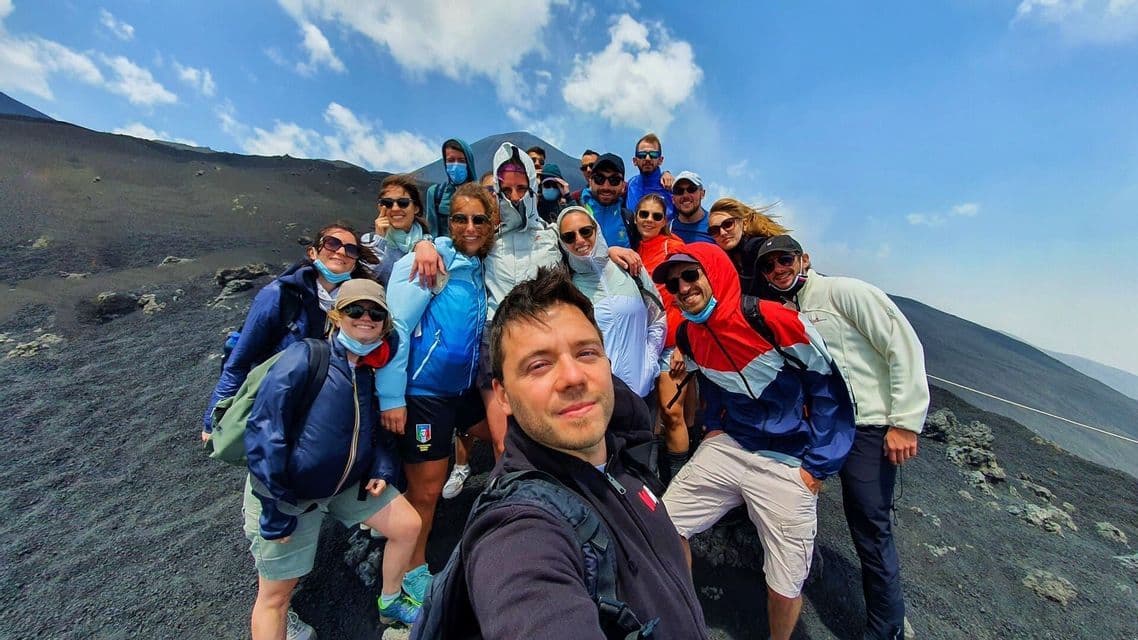 A WeRoad group trip posing for a selfie on the steep, dark soil of a volcanic mountain under a blue sky.