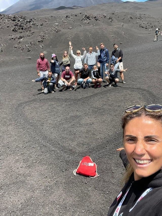 A woman takes a selfie with her WeRoad group trip as they pose on the dark, rocky soil of a volcanic landscape.