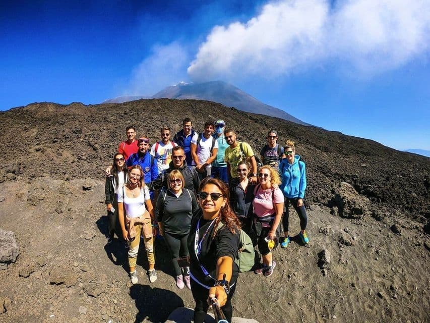 Un gruppo WeRoad si scatta un selfie su un fianco di montagna roccioso, con un vulcano fumante sullo sfondo sotto un cielo azzurro.