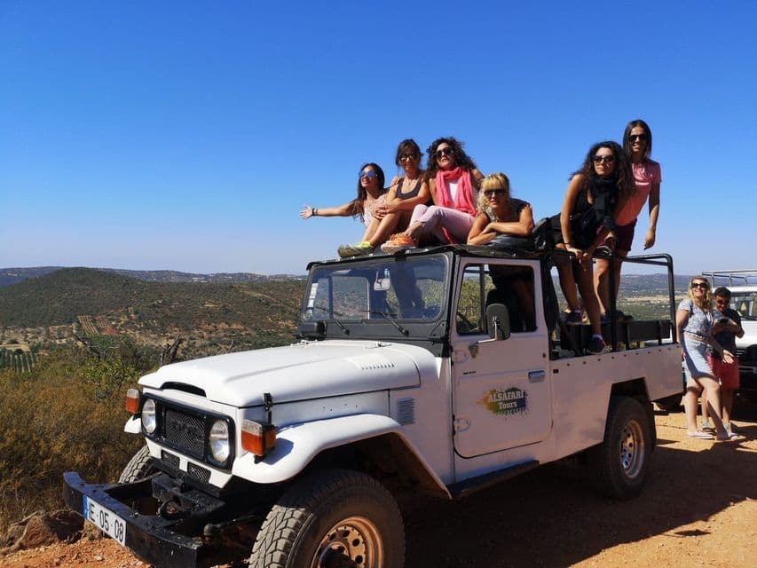 Eine WeRoad-Frauengruppe posiert auf einem weißen Safari-Jeep auf einer Schotterstraße, vor einer hügeligen Landschaft unter strahlend blauem Himmel.