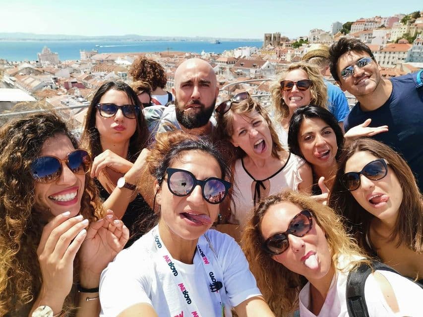 Eine WeRoad-Gruppe macht an einem sonnigen Tag mit Blick auf eine Stadt und das Meer ein verspieltes Selfie, bei dem sie die Zungen herausstrecken.