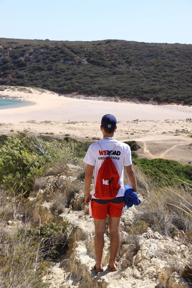 Un tour leader WeRoad, visto da dietro, si trova su un sentiero roccioso che si affaccia su una spiaggia sabbiosa e una collina verdeggiante.