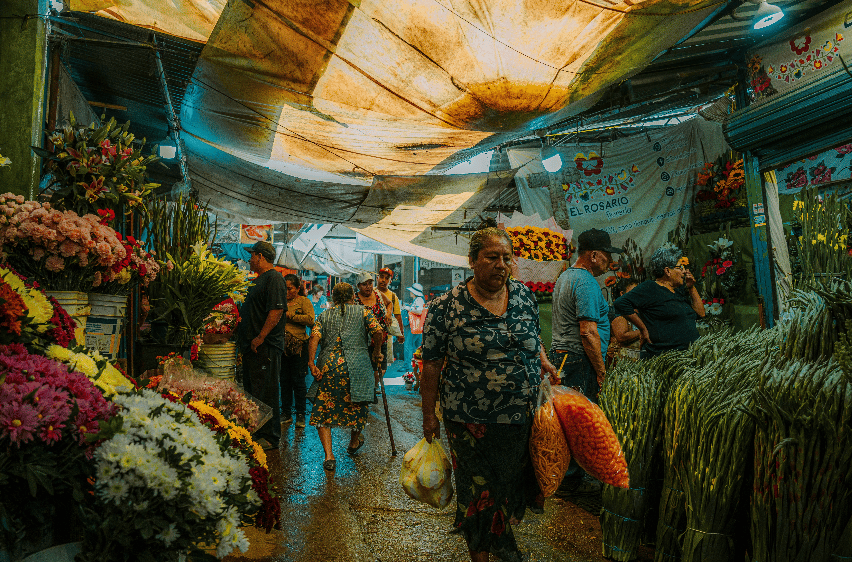 Menschen gehen auf einem nassen Weg durch einen belebten, überdachten Blumenmarkt, umgeben von Ständen voller farbenfroher Blumensträuße.