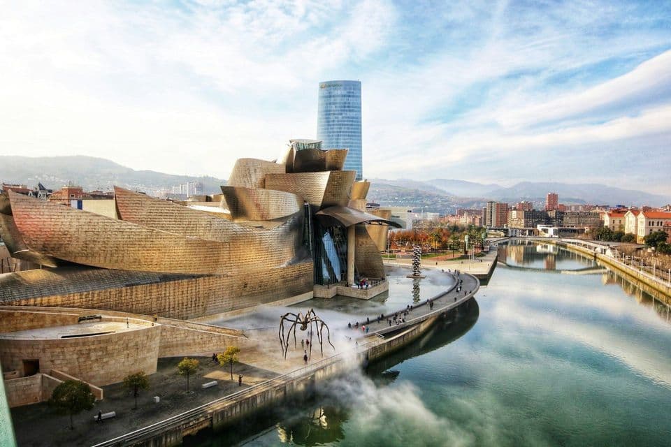 Un edificio metallico astratto con una scultura di ragno gigante sorge lungo un fiume, con la città e le colline visibili sullo sfondo.