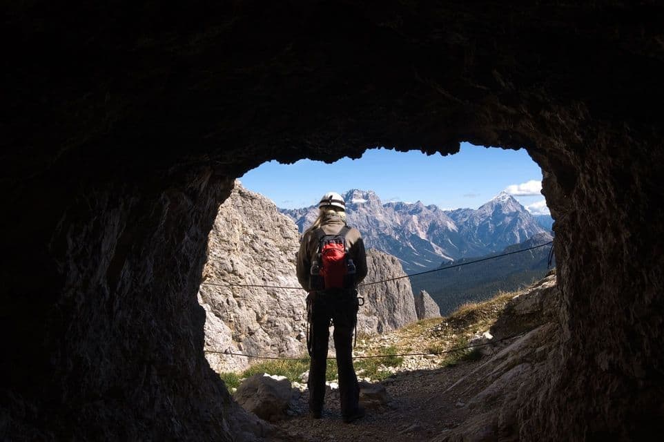 Un escursionista con un casco e uno zaino rosso si trova all'imboccatura di una grotta, guardando un'ampia catena montuosa illuminata dal sole.