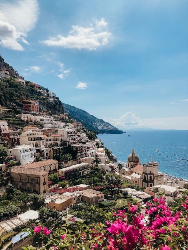 Un village bâti sur une falaise escarpée surplombant la mer bleue, avec des bougainvilliers roses au premier plan.