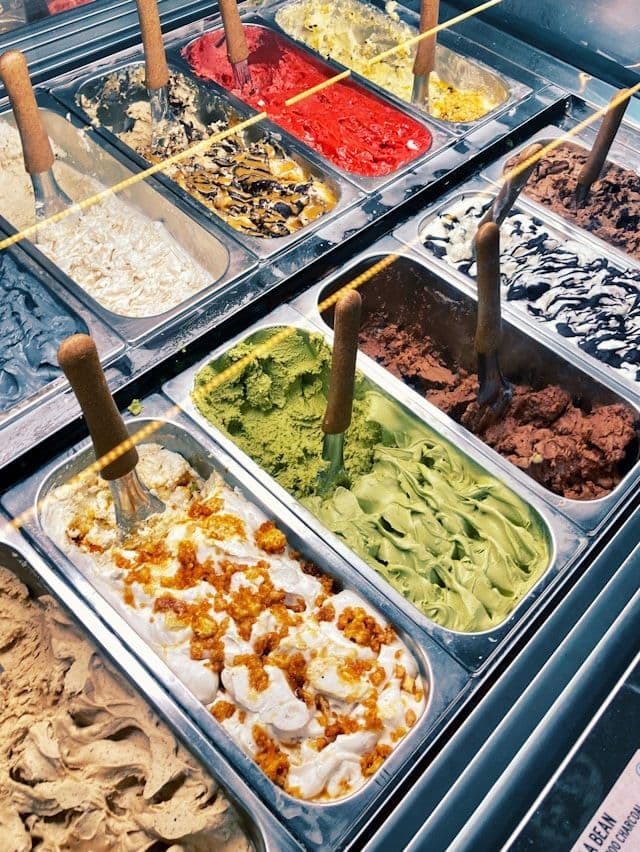 Una variedad de coloridos sabores de gelato dispuestos en cubas de metal dentro de una vitrina, cada uno con una cuchara para servir.