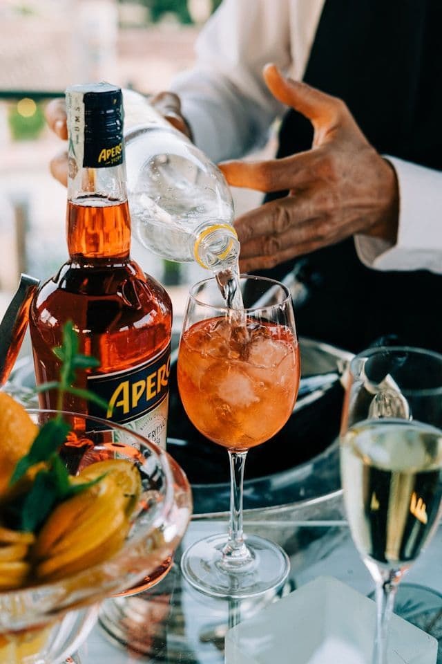 Les mains d'un barman versent un liquide transparent d'une bouteille dans un verre à vin avec de la glace, à côté d'une bouteille d'Aperol.