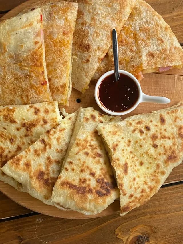 Triángulos de pan plano tostado, rellenos y cortados, presentados en una tabla de madera con un pequeño tazón de salsa oscura para mojar.