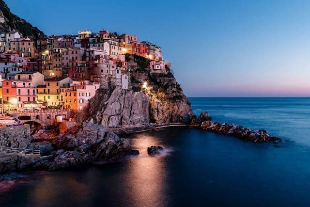 Colorful houses of a coastal village are illuminated on a rocky cliff overlooking the sea at dusk.