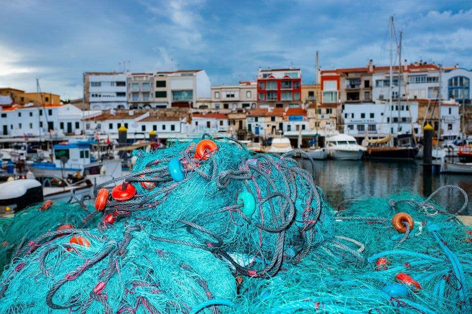 Una pila di reti da pesca blu brillante si trova su un molo, con barche ed edifici di una città portuale sullo sfondo.