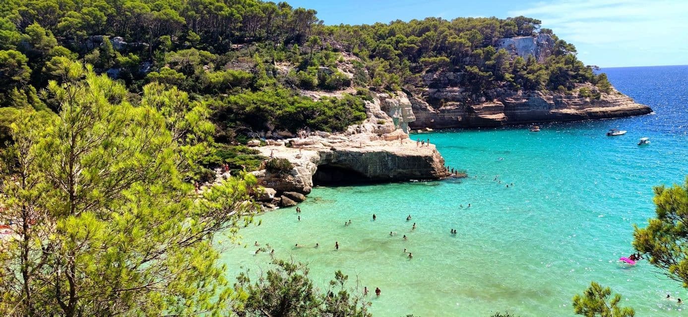 Vista dall'alto di persone che nuotano in una cala dalle acque turchesi, delimitata da scogliere rocciose con una grotta e alberi rigogliosi.