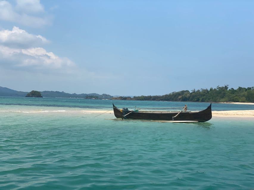 Una barca di legno ormeggiata su un banco di sabbia bianca circondato da acqua turchese, con isole coperte di alberi sullo sfondo sotto un cielo blu.
