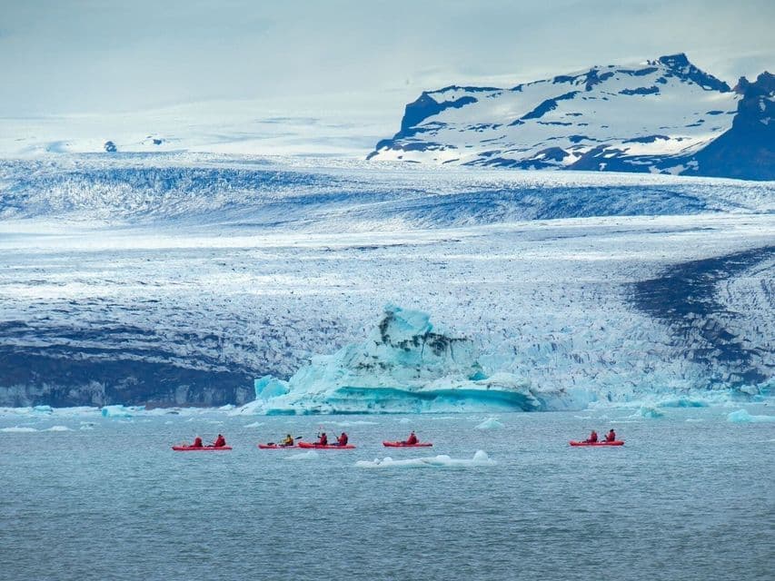 Un viaje en grupo de WeRoad rema en kayaks rojos por aguas heladas frente a un enorme glaciar y montañas nevadas.