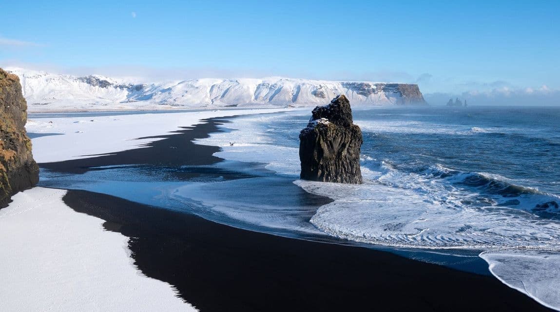 Un grande faraglione si erge nell'acqua al largo di una spiaggia di sabbia nera parzialmente coperta di neve, con montagne innevate sullo sfondo.