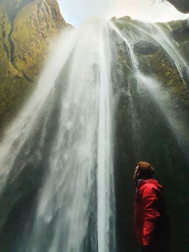 Une personne en veste rouge se tient au pied d'une cascade imposante, levant les yeux vers l'eau qui dévale le long d'une falaise moussue.
