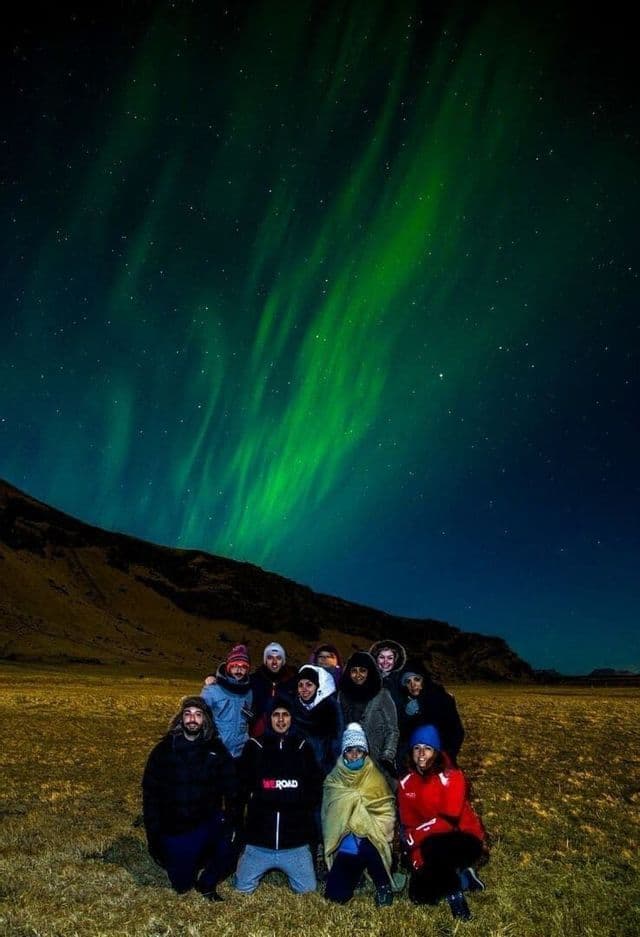 A WeRoad group trip posing for a photo in a field at night under the green aurora borealis and a starry sky.