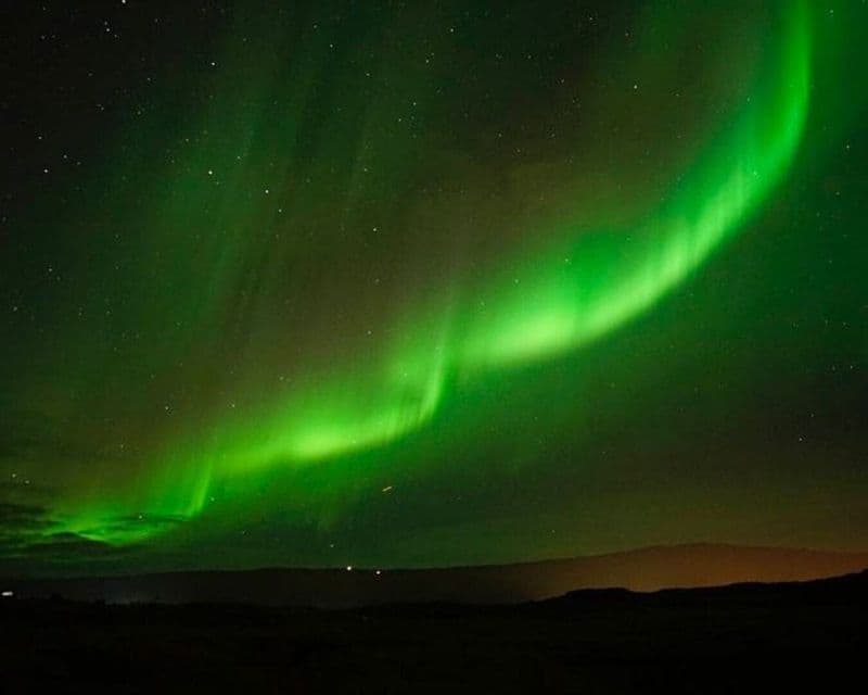 Un'aurora boreale di un verde brillante si inarca in un cielo notturno stellato sopra la silhouette di un paesaggio scuro.