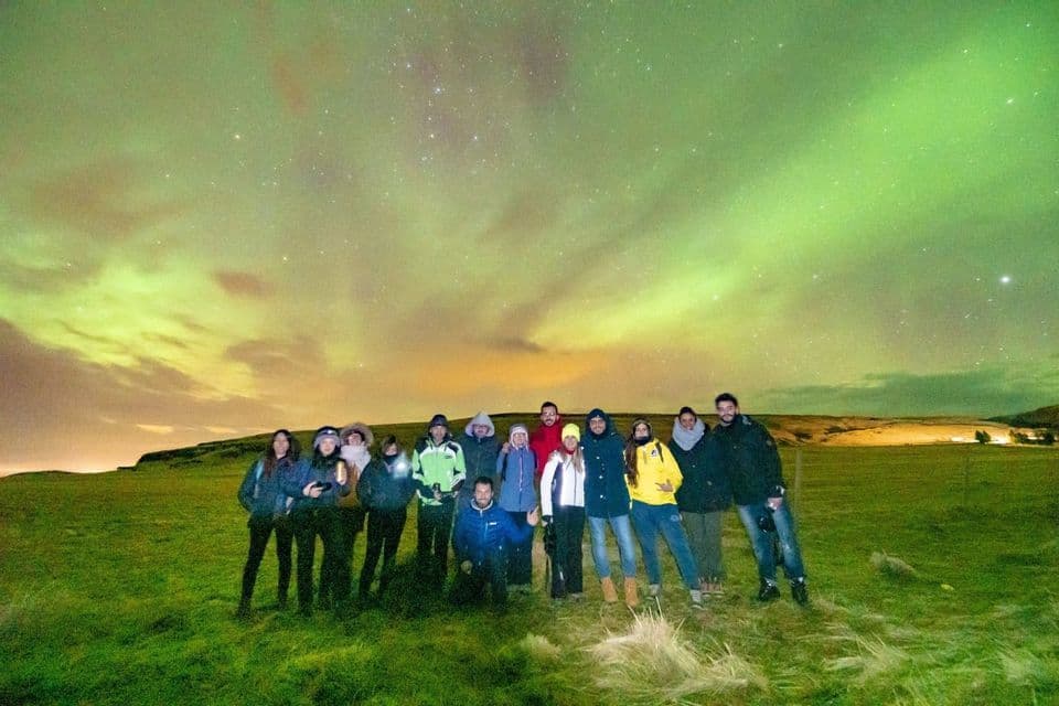 Un groupe WeRoad en voyage posant pour une photo sur une colline herbeuse, sous les aurores boréales vertes et un ciel étoilé.