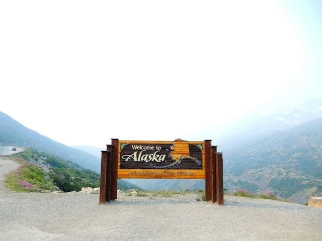 The wooden 'Welcome to Alaska' sign stands on a gravel overlook with a hazy mountain range in the background.
