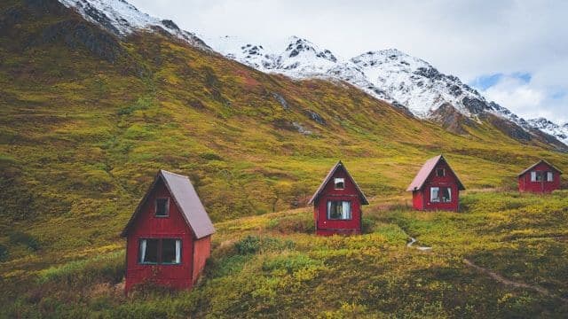 Four small red cabins sit on a green and yellow hillside at the base of a snow-capped mountain.