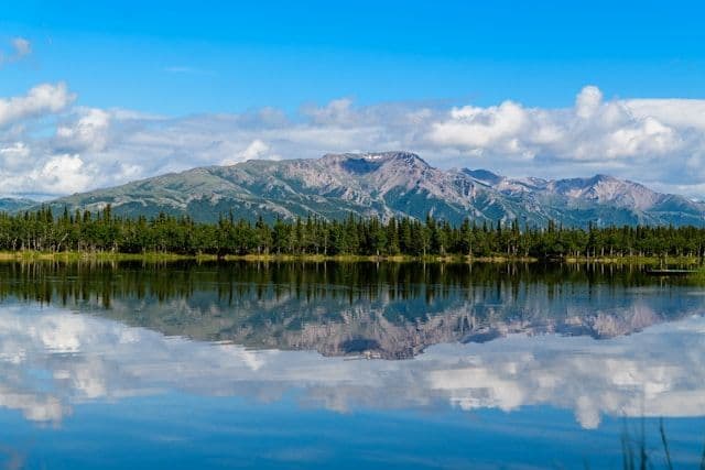 A mountain range and a line of trees reflecting on the calm surface of a lake under a partly cloudy sky.