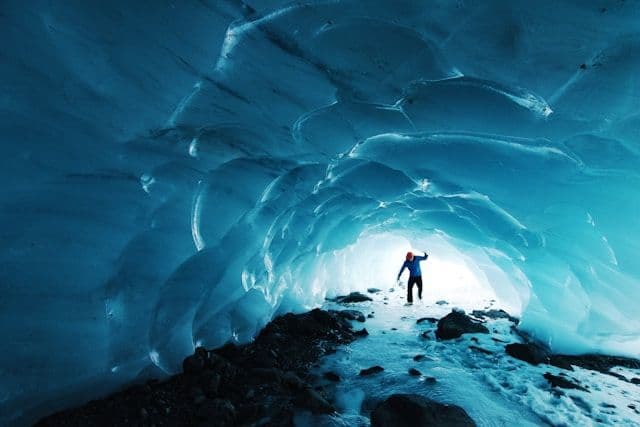 A person in a blue jacket and red beanie stands inside a glowing blue ice cave on a snowy path.