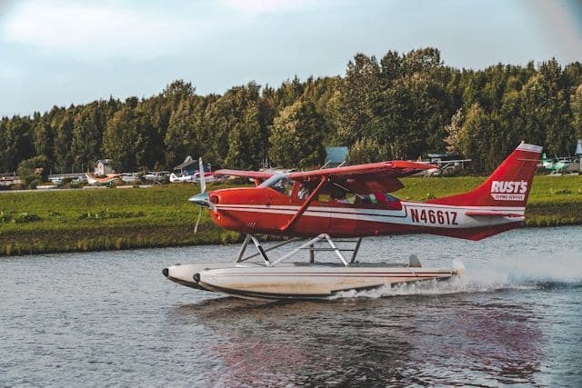 A red and white seaplane taxis on the water, creating a wake, with a green forested shoreline in the background.