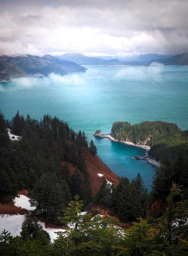 An elevated view of a turquoise bay surrounded by pine-covered mountains and patches of snow under a cloudy sky.