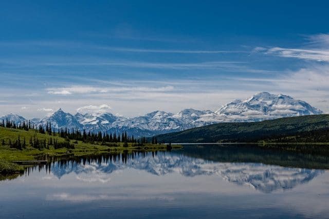 A range of snow-capped mountains and a pine forest reflect perfectly in the still water of a lake under a blue sky.