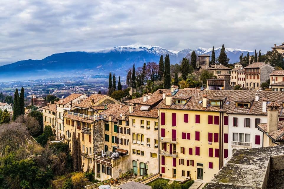Panoramica sui tetti in terracotta di una città colorata su una collina, con una valle e montagne innevate sullo sfondo.