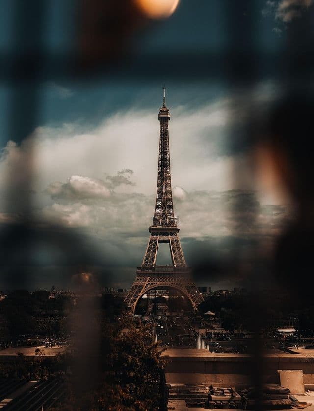 A tall, iron lattice tower stands in the center of a cityscape under a dramatic, cloudy sky, viewed through a blurry foreground.