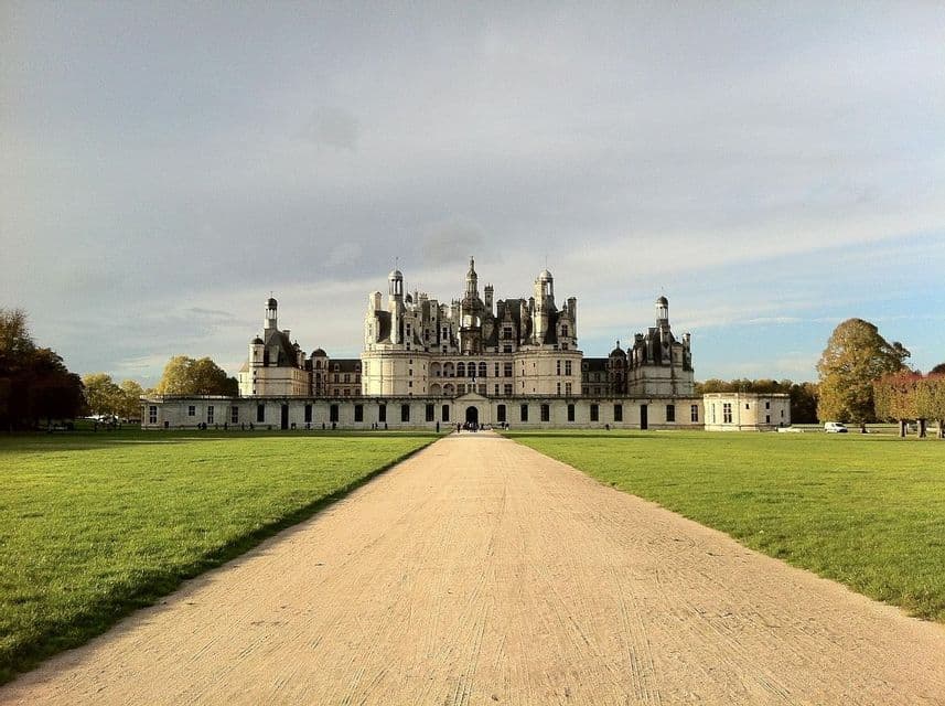 A straight gravel path cuts through a vast green lawn, leading towards a large, ornate white chateau.