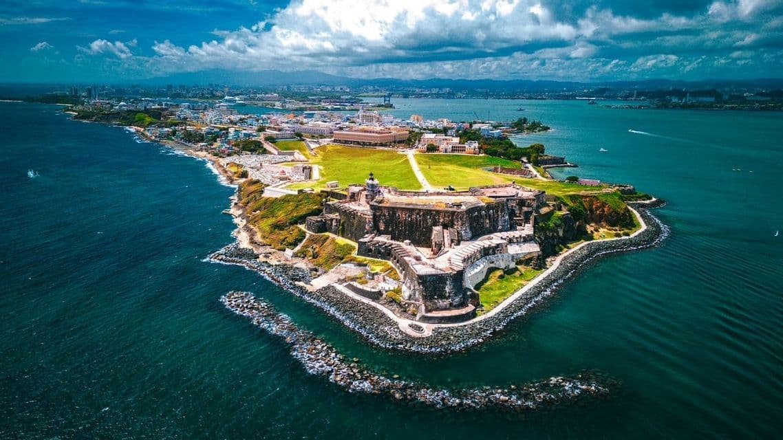 An aerial view of a historic stone fortress on a green peninsula surrounded by deep blue water, with a city skyline in the background.