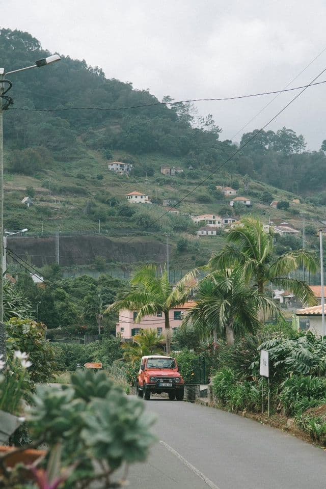 A red off-road vehicle on a narrow road surrounded by lush greenery, with houses scattered across a forested hillside in the background.