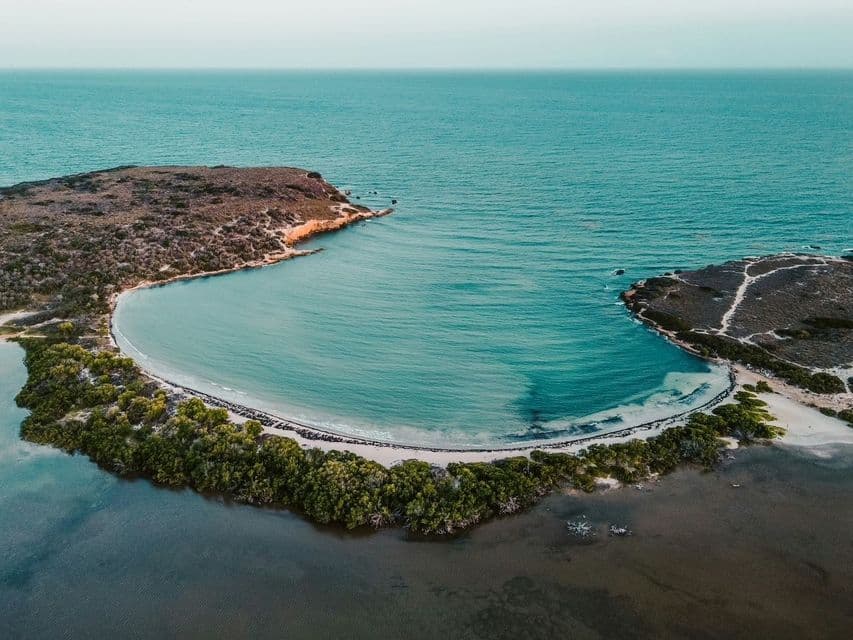 An aerial view of a crescent-shaped bay with a white sand beach, bordered by green mangroves and opening into the turquoise sea.
