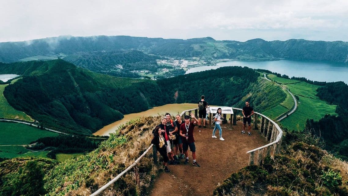 A WeRoad group trip poses for a photo on a scenic viewpoint overlooking a valley with two lakes and lush green hills.