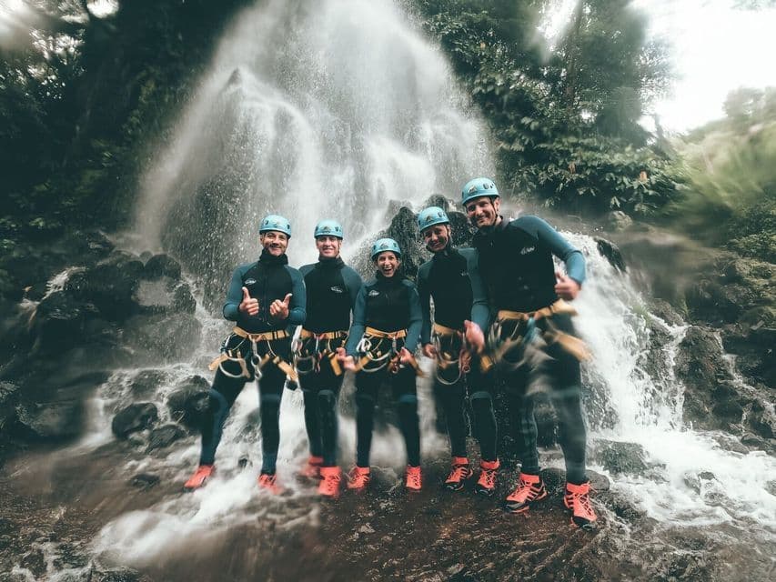 A WeRoad group trip in wetsuits and helmets posing with thumbs-up gestures in front of a cascading waterfall.
