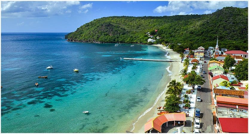 Vue aérienne d'une ville côtière tropicale avec une plage de sable, une longue jetée et des bateaux flottant dans une eau turquoise claire.