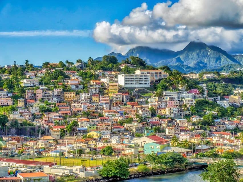 Des maisons colorées et des arbres luxuriants garnissent une ville à flanc de colline, dominée par de grandes montagnes sous un ciel partiellement nuageux.