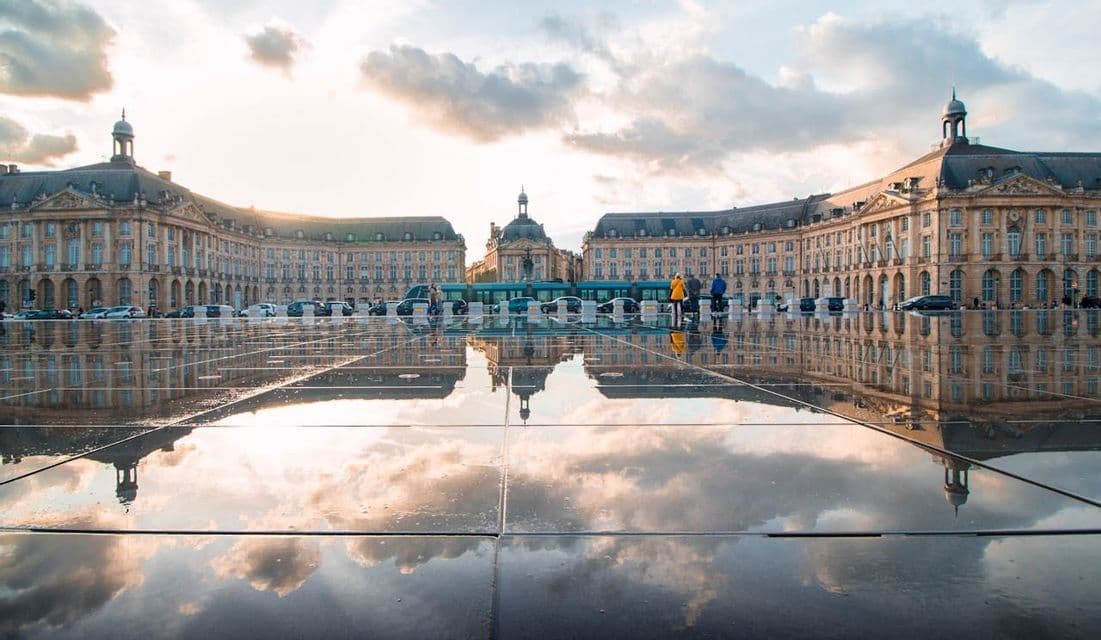 A symmetrical historical building and a cloudy sky reflecting on the wet pavement of a large public square at dusk.