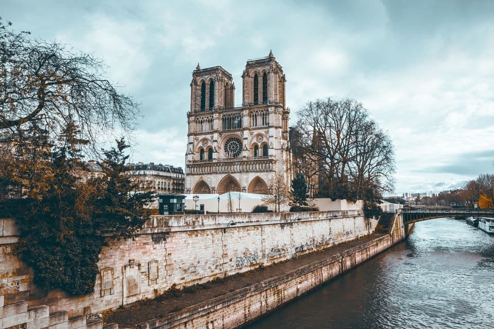 A Gothic cathedral with two bell towers and a rose window, seen from across a river on a cloudy day with bare trees nearby.