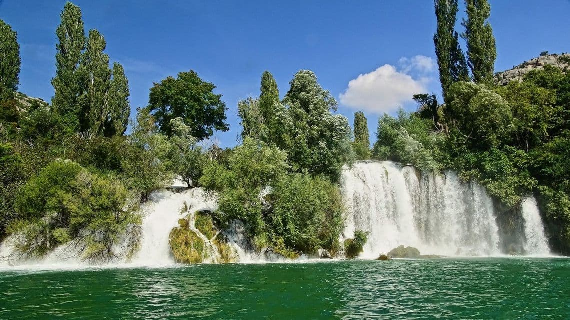Una vasta cascata si riversa sulle rocce in un fiume verde smeraldo, circondata da alberi lussureggianti sotto un cielo azzurro e limpido.