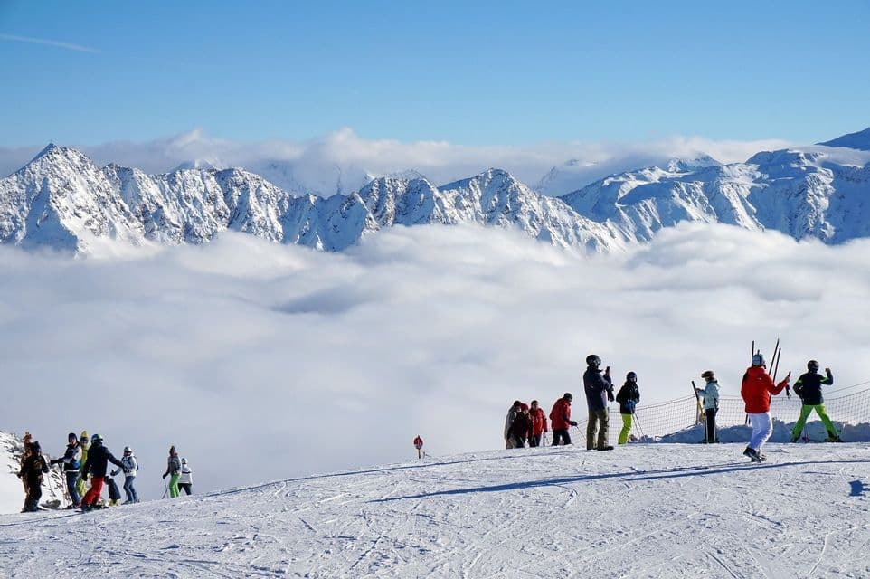 Viaje en grupo WeRoad de esquiadores en la cima de una montaña nevada, observando picos sobre un mar de nubes bajo un cielo azul.