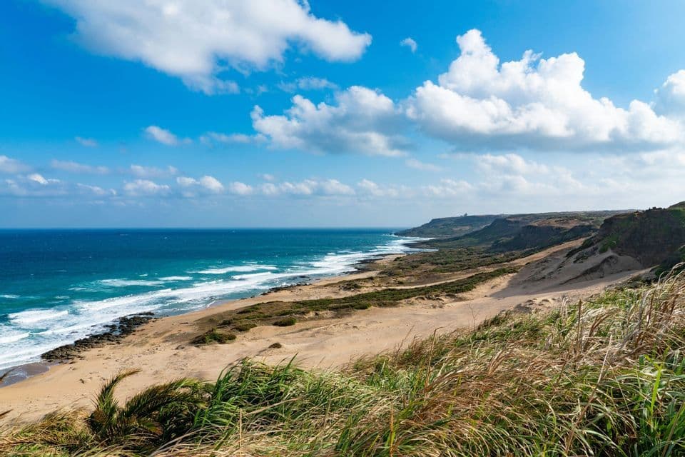 Ein malerischer Blick auf eine sandige Küstenlinie mit blauen Meereswellen von einer grasbewachsenen Anhöhe unter einem teils bewölkten Himmel.
