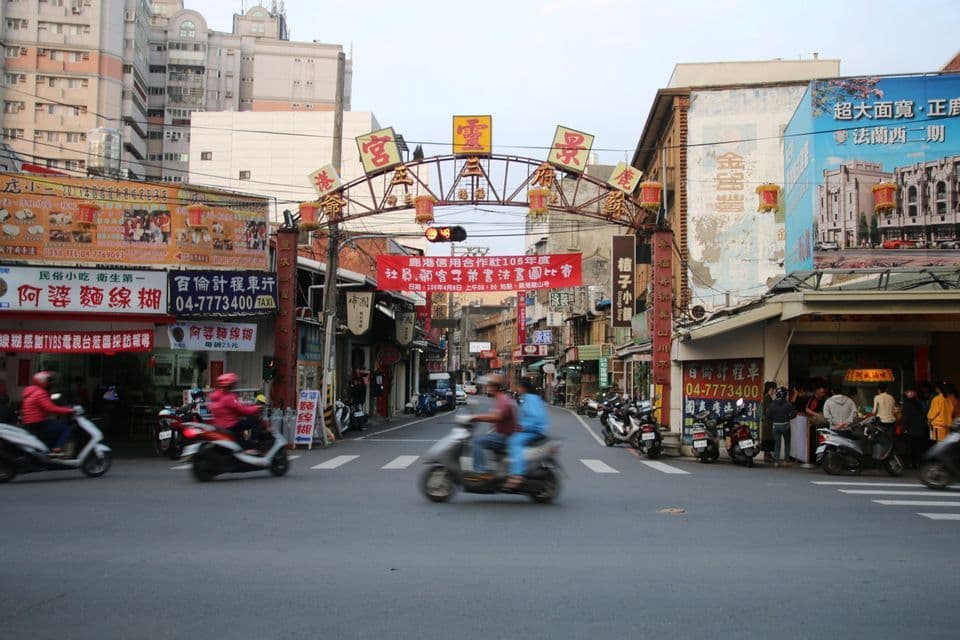 Rollerfahrer befahren eine belebte Stadtstraße und passieren dabei einen traditionellen Torbogen, geschmückt mit Laternen und chinesischen Schriftzeichen.